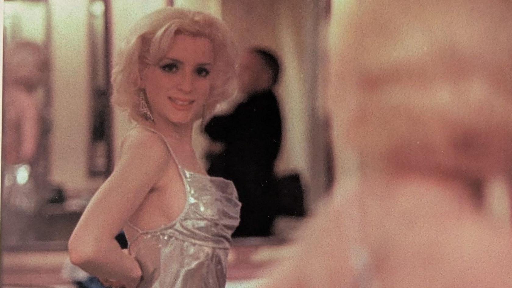 Glamorous young woman looks at herself in the mirror. She has short blond curly hair, a shiny silver dress and long diamond style earrings. 