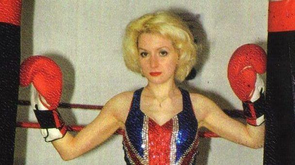 Young blond woman with a stern expression, she has curly short hair and wears a glittery union jack dress. She has red boxing gloves with her hands raised to her shoulders. 