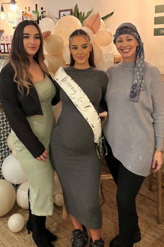 Three women stood in a party setting. The woman on the left is brunette, wearing a green dress with black boots and a black cardigan. The woman in the middle is pregnant, wearing a grey dress and a satin sash. The woman on the right is smiling, wearing a grey jumper and black trousers. She is also wearing a grey scarf on her head. 