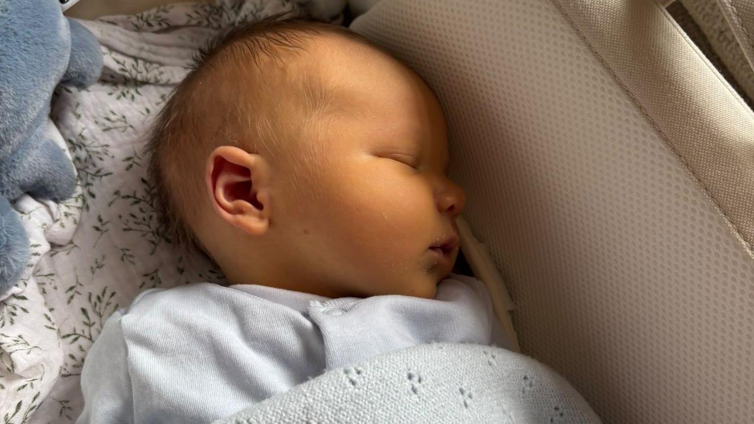 Baby Gwilym sleeping in his crib. He's a newborn and wearing a baby blue suit. He's wrapped in a blue blanket and lying on a floral sheet with a soft toy at the top of the photograph.