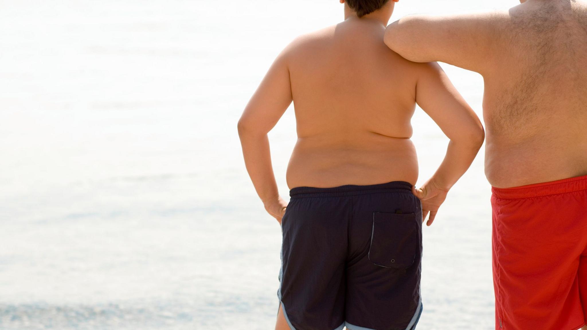 A boy and his father stand in their swimming trunks looking at the sea. Both are overweight or obese