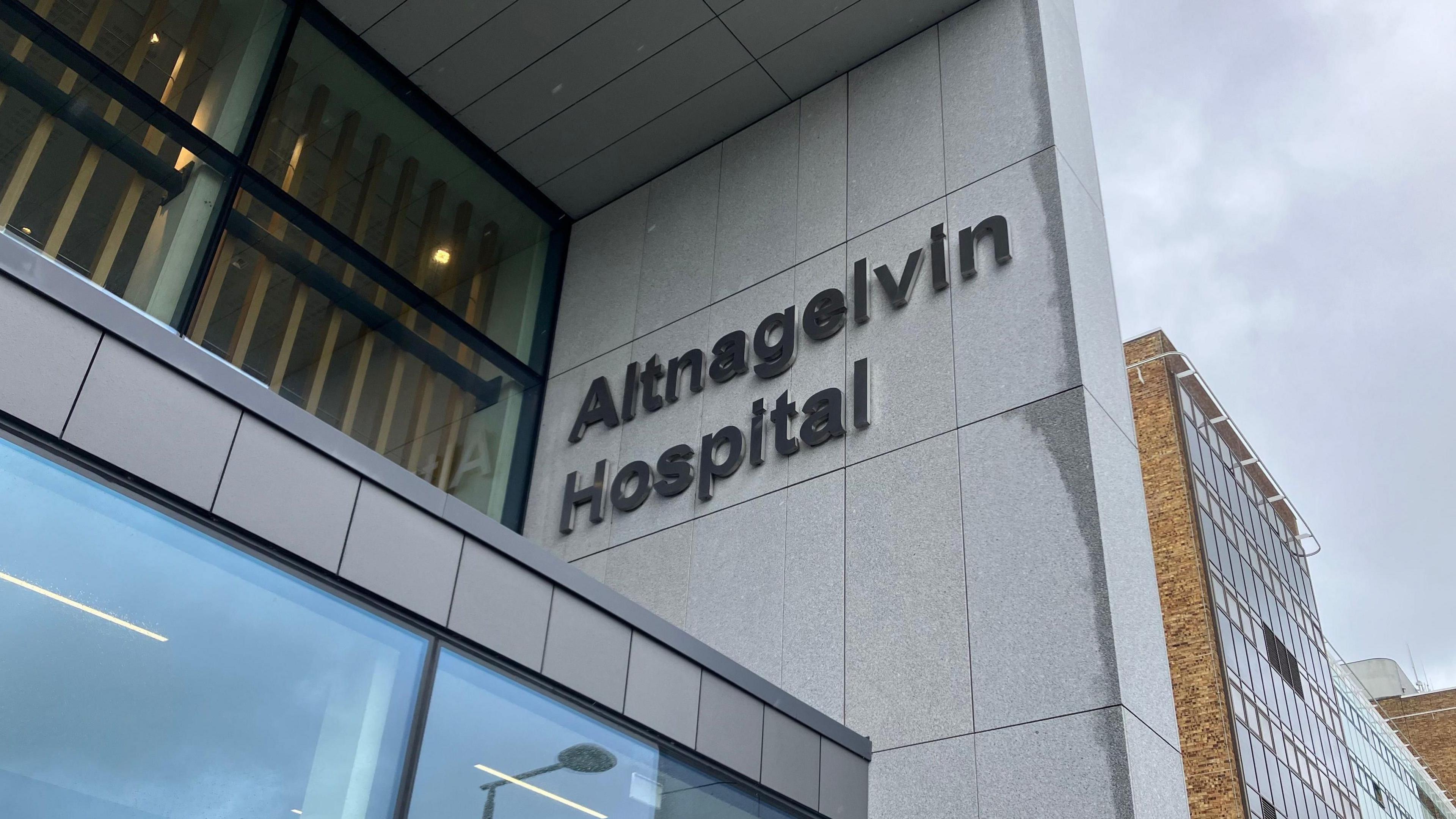 A close up of a sign outside Altnagelvin Hospital, which says the name of the building in large lettering. It is a tall building with a mix of facades including brown brick and grey cladding.