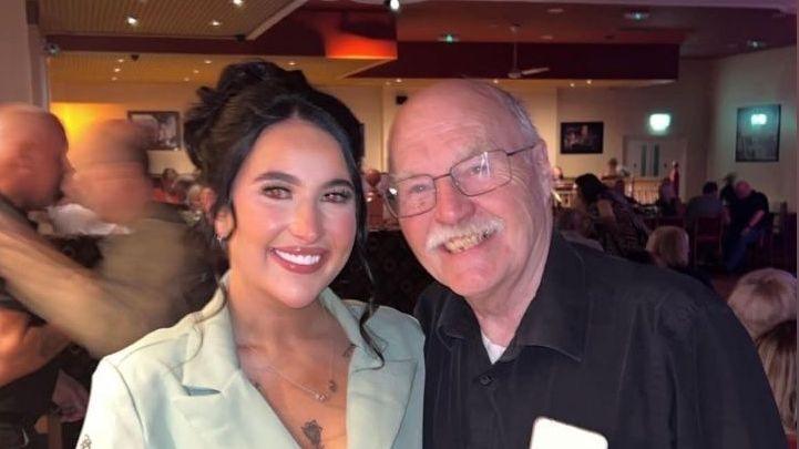 A woman with dark hair, wearing a green jacket, stands beside her grandfather. He is wearing a black shirt and black trousers. They are in a restaurant.