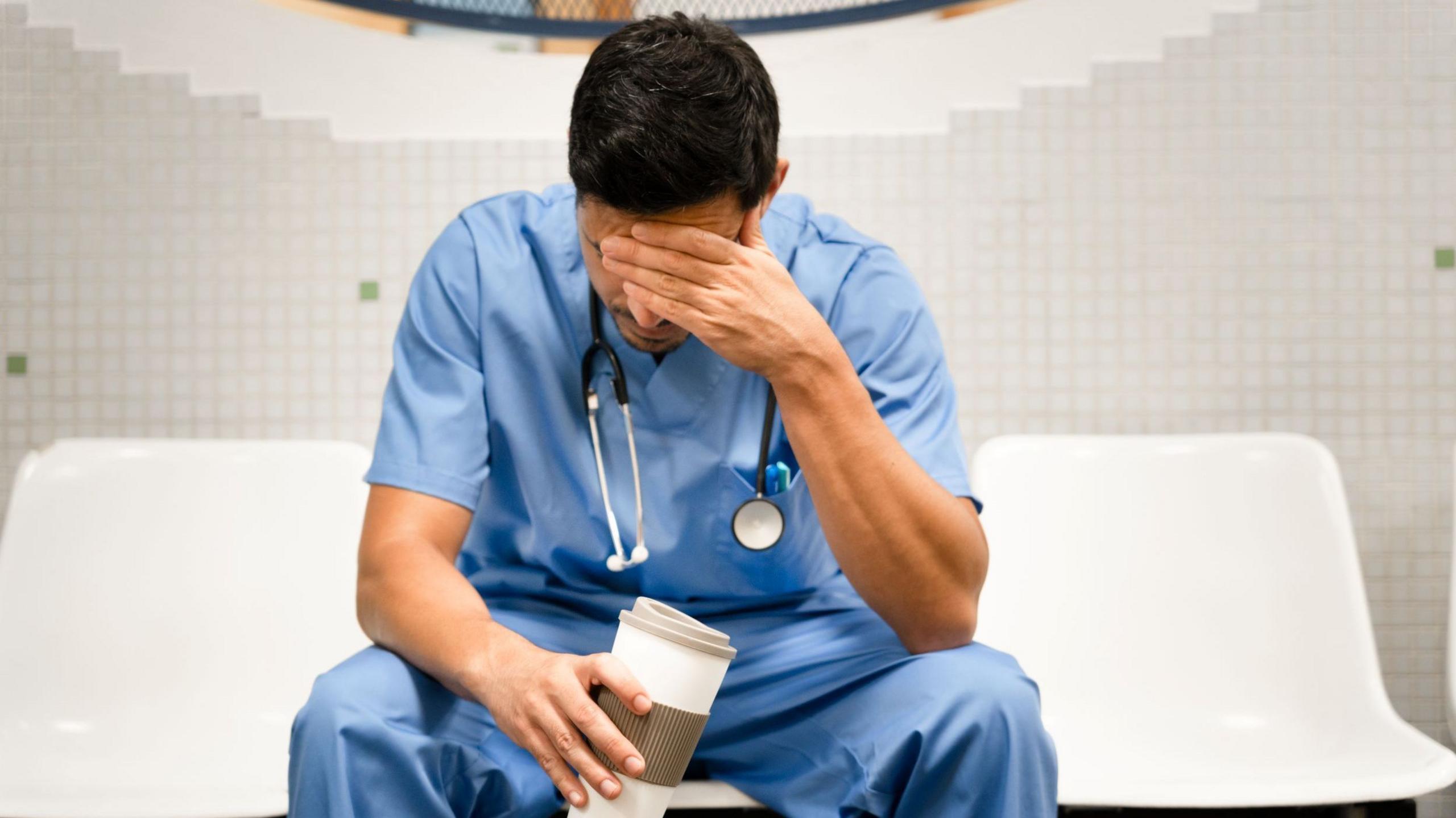 A male doctor in blue scrubs and short dark hair is sitting on a while plastic chair. He is leaning his elbows on his knees and has his head bowed with his left hand held over his face. In his right hand is a coffee cup. There's a stethoscope around his neck.