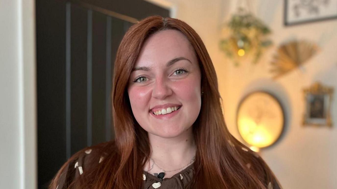 Amy-Jane is stood in her living room and is smiling at the camera. She has long reddish-brown, straight hair and is wearing a brown and white spotty blouse. 