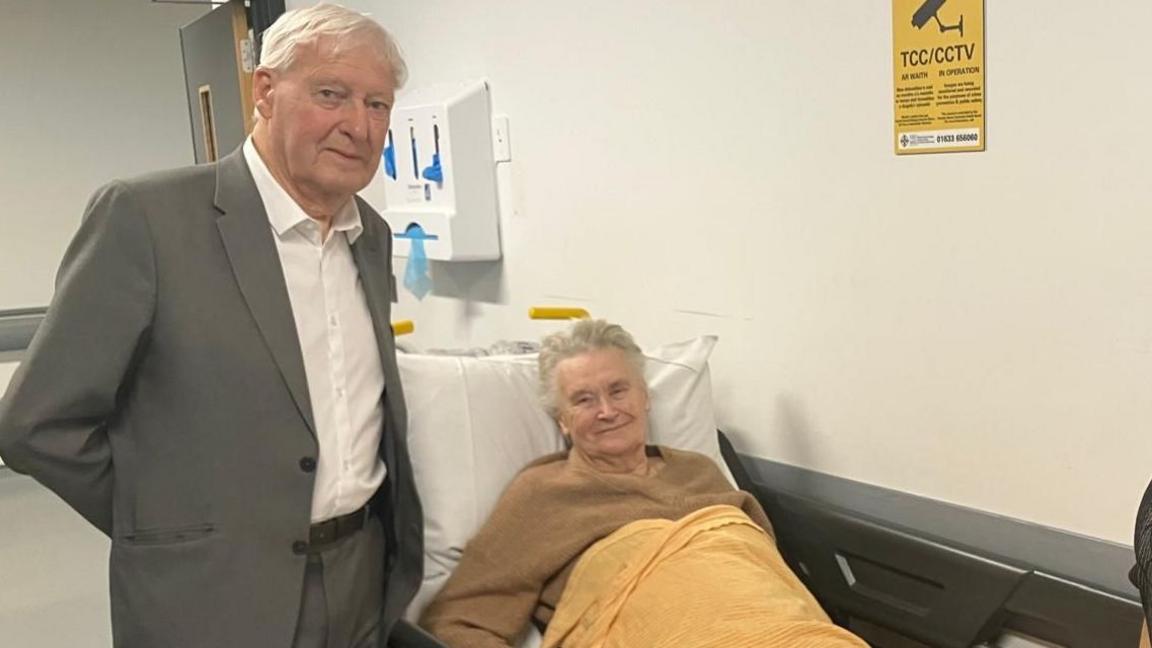 An elderly woman with short hair is seen propped up on a hospital bed. She wears a camel coloured jumper and her lower half is covered by a yellow blanket. Her husband also with cropped white hair stands to her right with his arms behind his back. He is wearing a white shirt and a grey suit trousers and jacket.