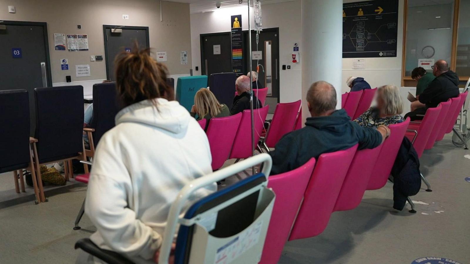 Two rows of pink chairs have a number of patients in them. The closes row sees the backs of 5 people one is hooked to an IV drip. The closest patient wears a hoodie and is sat in a hospital wheelchair. In front of them there high back blue chairs - only the outlines of two patients can be made out.
