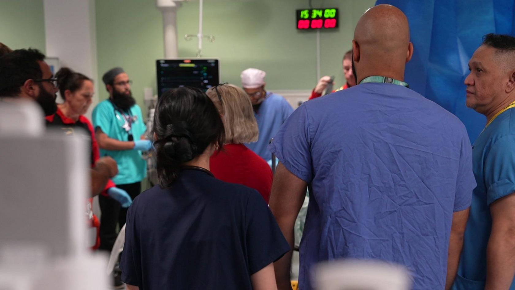 A number of doctors and nurses crowd around a trolley. They wear light blue, navy and blue scrubs. Medical equipment can be seen out of focus in the foreground. 