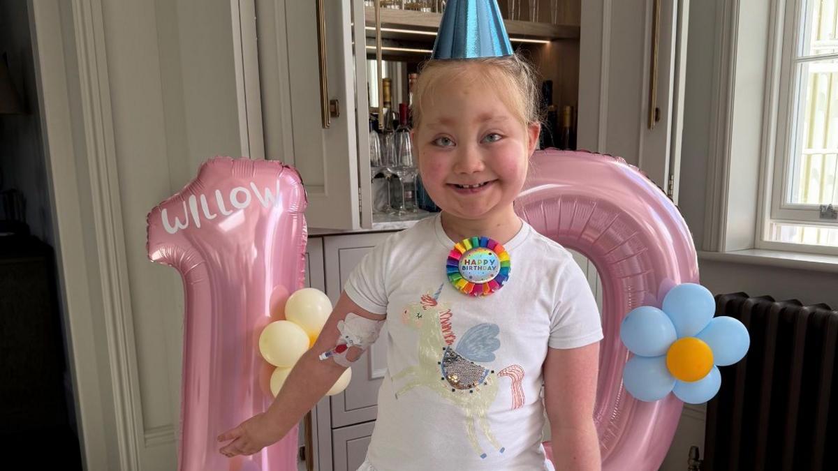 Willow stands in front of pink number 10 balloons. She has a happy birthday badge on her unicorn T-Shirt and is wearing a blue party hat