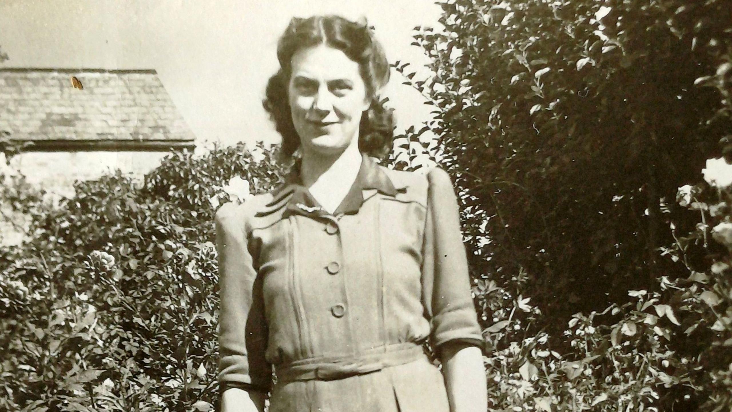 A black and white photo of a woman smiling at the camera in front of bushes