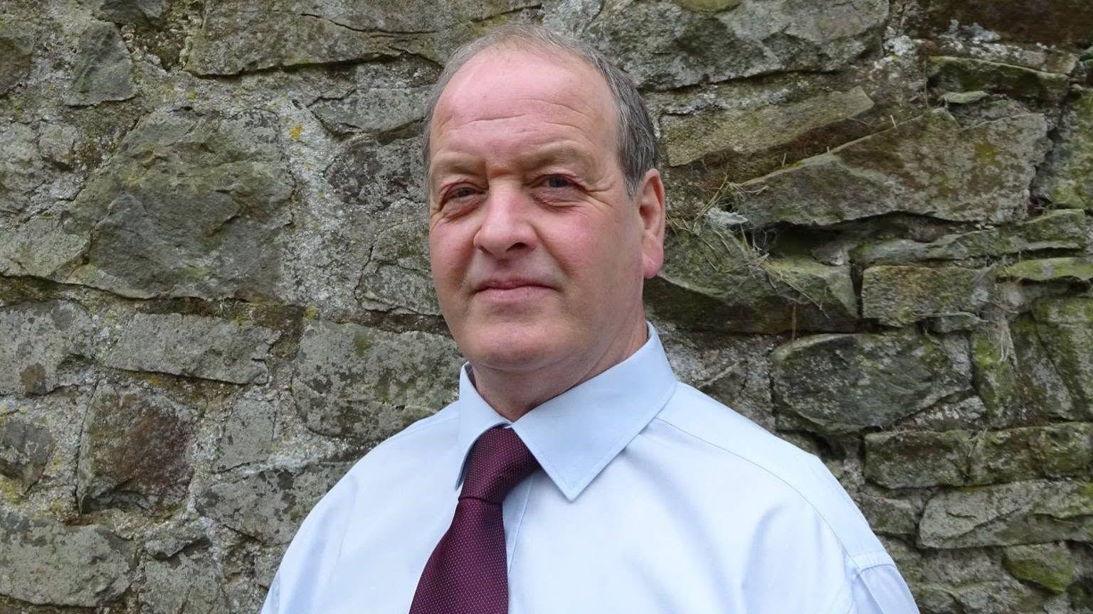 Alan Watson wearing a burgundy tie and a light blue shirt. He stands in front of a stone wall and looks at the camera. He has grey short hair and blue eyes.