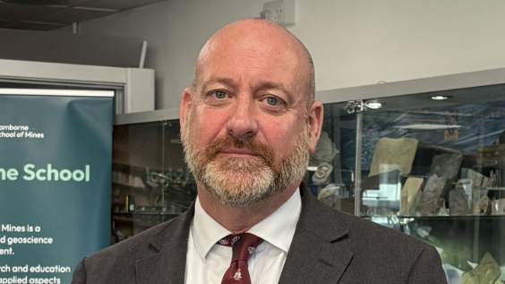 Head and shoulders shot of Perran Moon, who has a neat beard and is wearing a dark brown suit with a white shirt and burgundy tie.