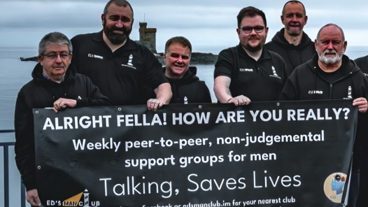 A group of men in black tops standing on Douglas Promenade holding a banner about the support services that says talking saves lives.
