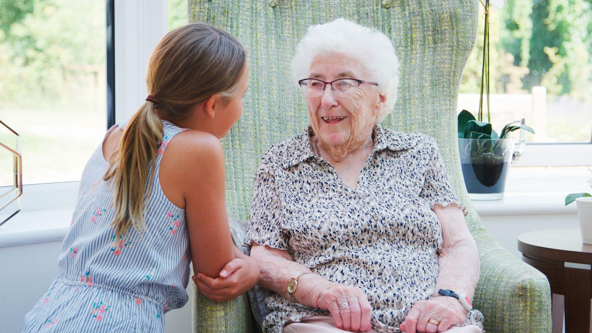 A young girl talking to an elderly woman. The girl has long, fair hair tied back in a ponytail and is wearing a blue and white striped jumpsuit with a pink floral motif. The woman, who is sitting in a green fabric armchair, has short, white hair, glasses and is wearing a beige, white and black patterned blouse with short sleeves.