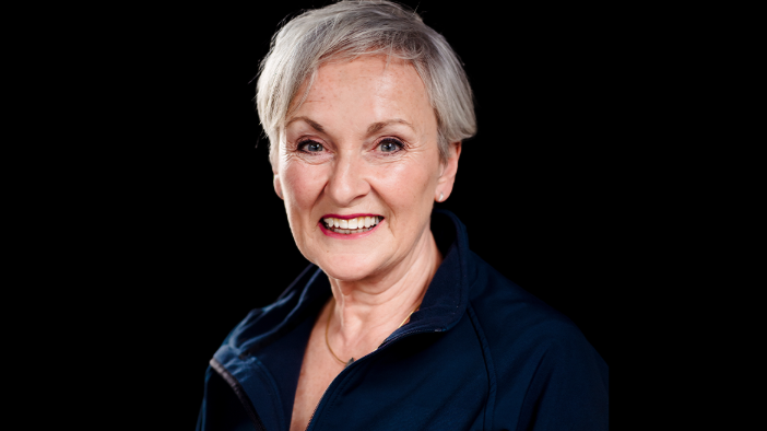 Siobhan Casey - a woman with short, grey hair - smiles at the camera during a photo shoot. She is wearing a dark blue jacket and is pictured against a plain black background.