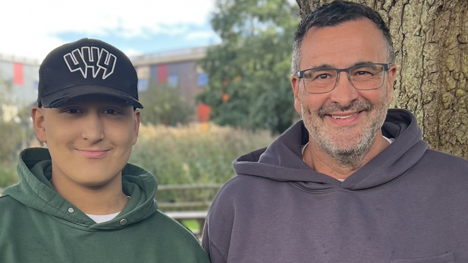 Ethan on the left and Lloyd on the right look at the camera and smile. Both are hoodies standing side by side outdoors beside a large tree. Ethan is wearing a black baseball cap with a white logo and Lloyd r wearing glasses. Trees, reeds and a building are out of focus in the background.