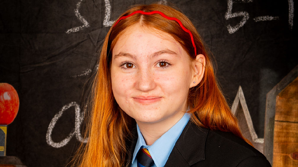 A girl sitting for a school photo. She has long red hair and a red head band on. She has a black blazer, a light blue shirt and a black, yellow and blue tie. She smiles at the camera.