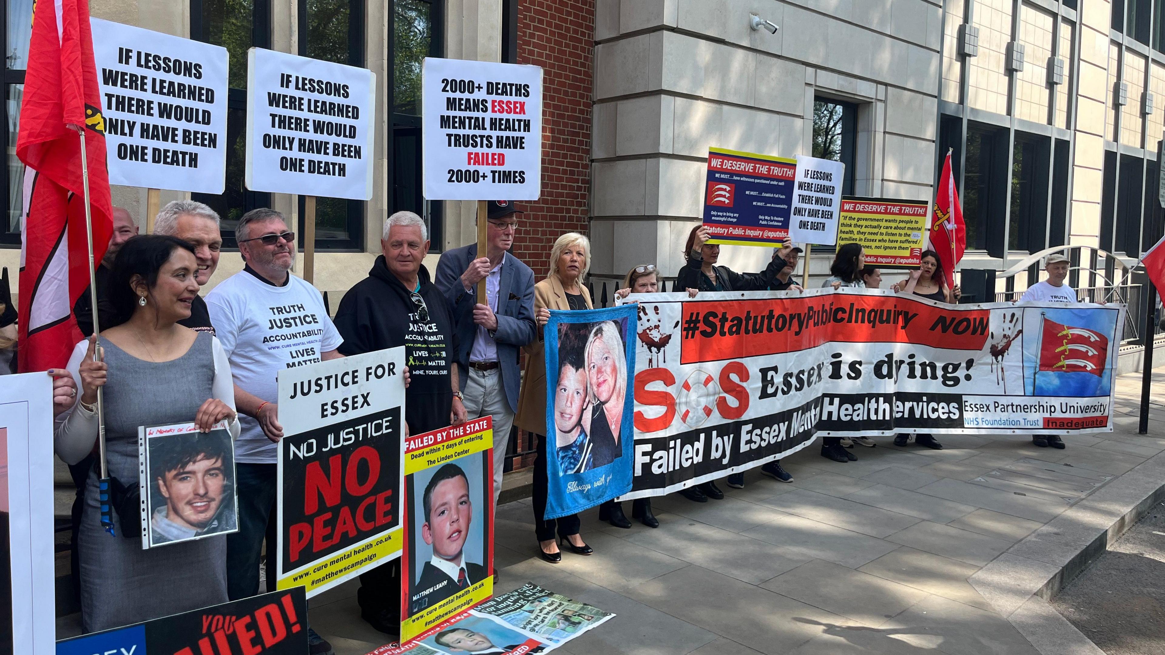 Several campaigners stand outside a building holding placards and signs calling for changes in how mental health services are handled. 