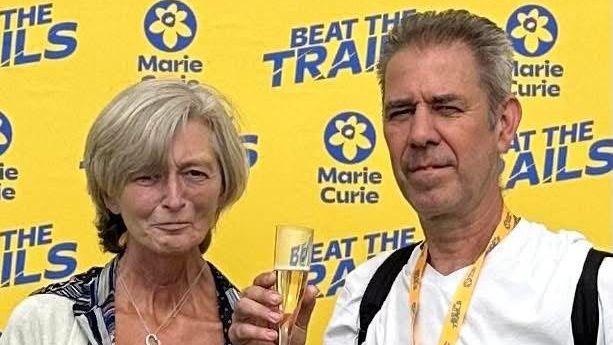 A woman stood next to a man who has completed a race for Marie Curie. He has a medal around his neck and holding a glass of bubbly. 