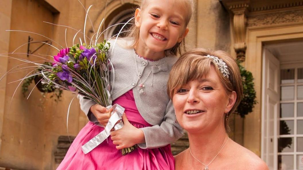 A mum and daughter at a wedding. The little girl with blonde hair is smiling. She's in a pink dress with a grey cardigan and holding a bunch of flowers. Her mum has a silver clip in her hair. 