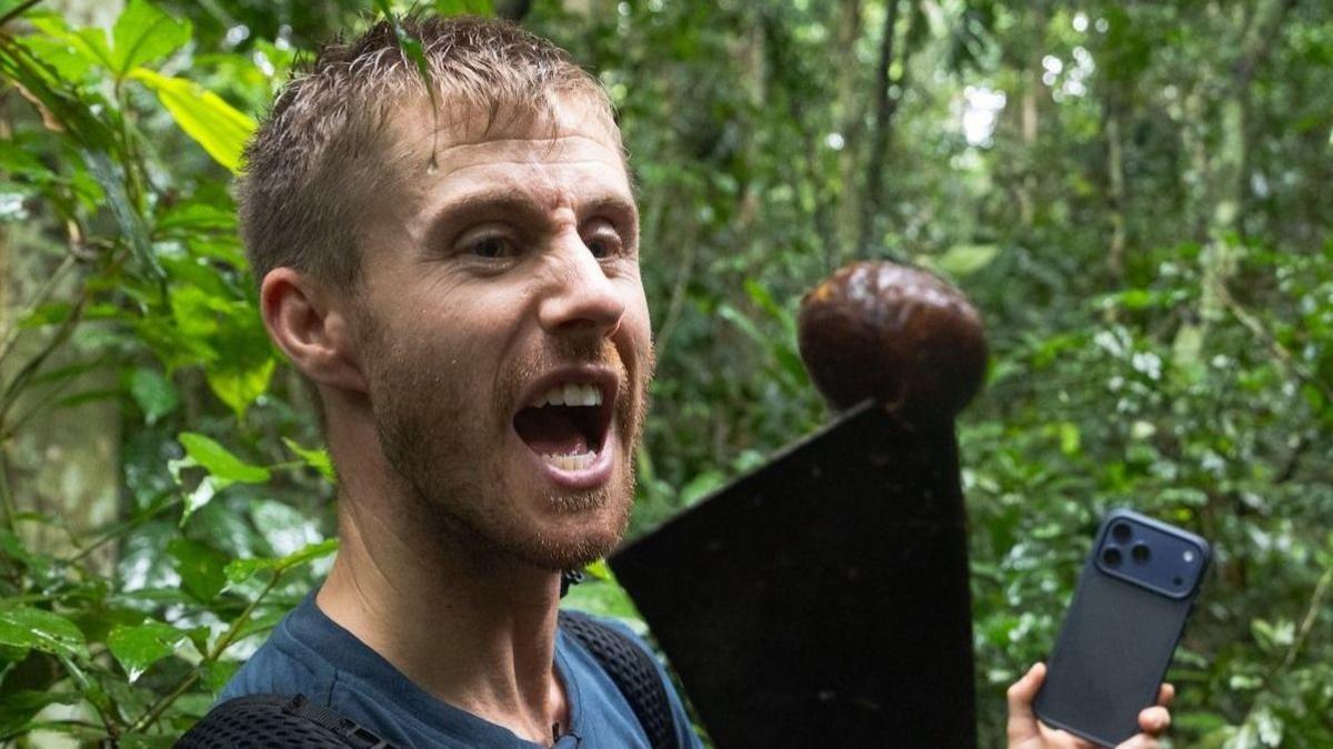A man holding a machete in the forest, in the other hand he holds his phone. He has a shouting expression.