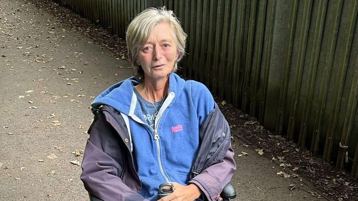 An ill woman sat in a wheelchair. She has short grey hair and wearing a Superdry zipped hoodie with a purple raincoat on top. 