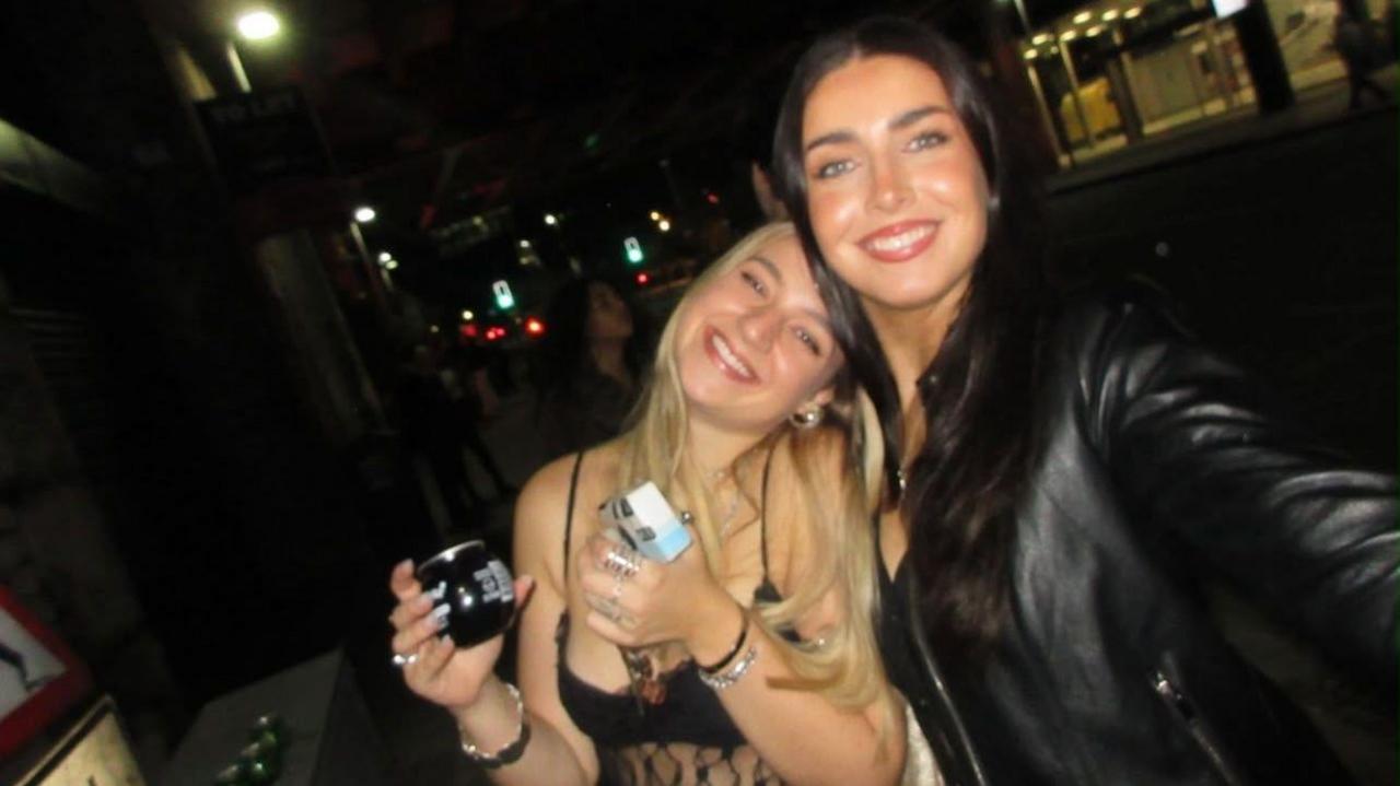 There are two girls in a dark bar smiling at the camera . On the left a blonde girl is wearing a strappy top and holding a drink. She leans into her friend on the left who is holding the camera to take a selfie of the two of them. The girl on the right has long brown hair and is wearing a black leather jacket.