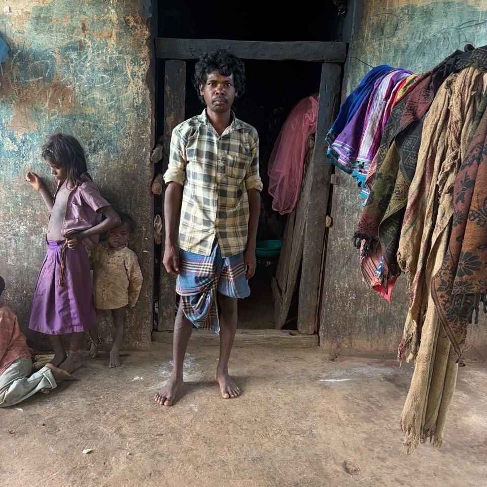 A man standing in the dark doorway of a sparse hut, with two children in the background