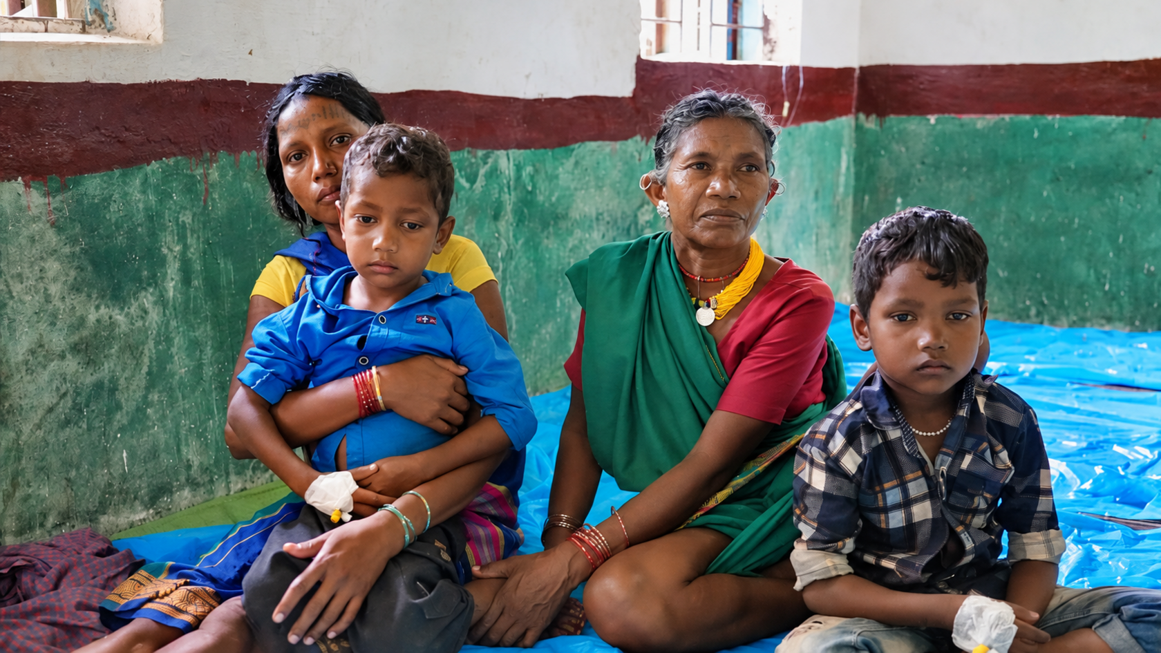 Two women and two children sitting on the floor of a medical camp in a village in India's Madhya Pradesh. The children have bandages on their hands where they were given a drip