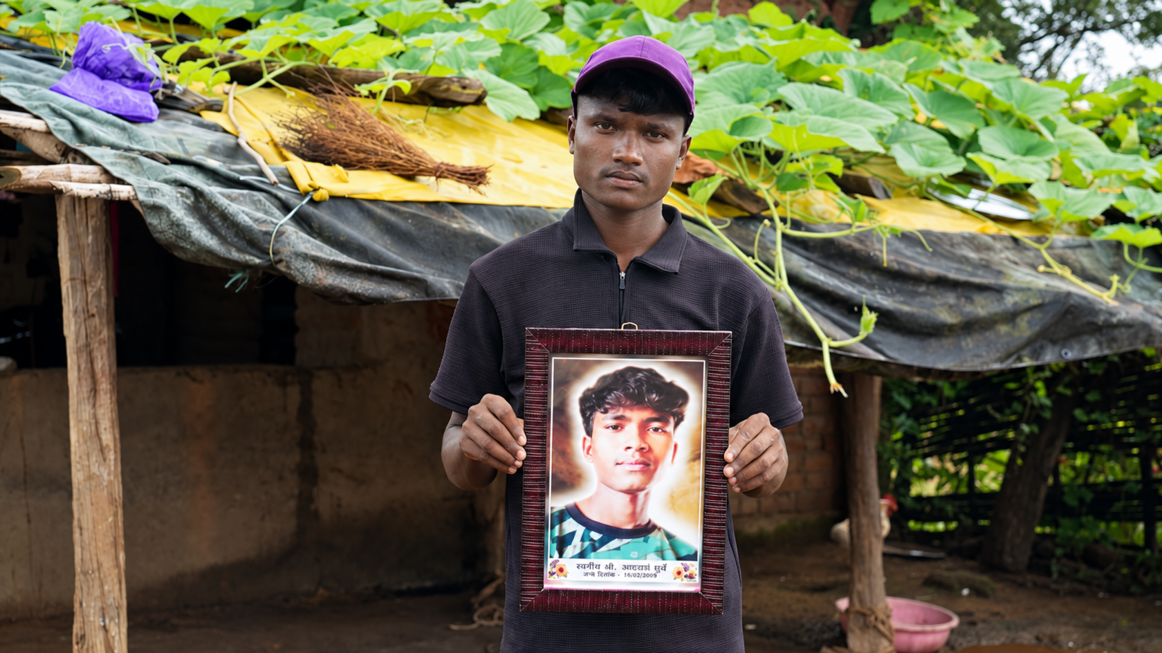A young man standing in front of a shed holds up a framed picture of a deceased teenage boy, which has the date of his death printed under his headshot.