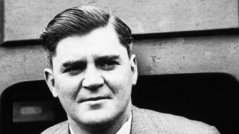 Black and white headshot of Aneurin Bevan taken in 1934. His dark hair is parted on the side and slicked back. A suit jacket and white shirt collar are visible. He appears to be leaning out of a train carriage door and is looking slightly to the right of the photo. He has a serious expression.