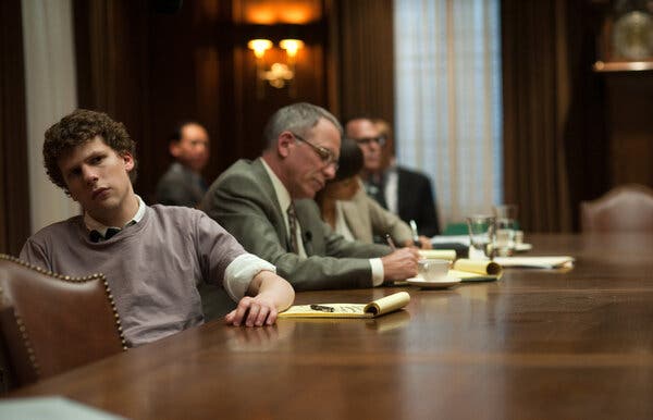 Jesse Eisenberg sitting at a board room table in a scene from “The Social Network.” He is wearing a sweater and collared shirt; others in suits sit nearby.