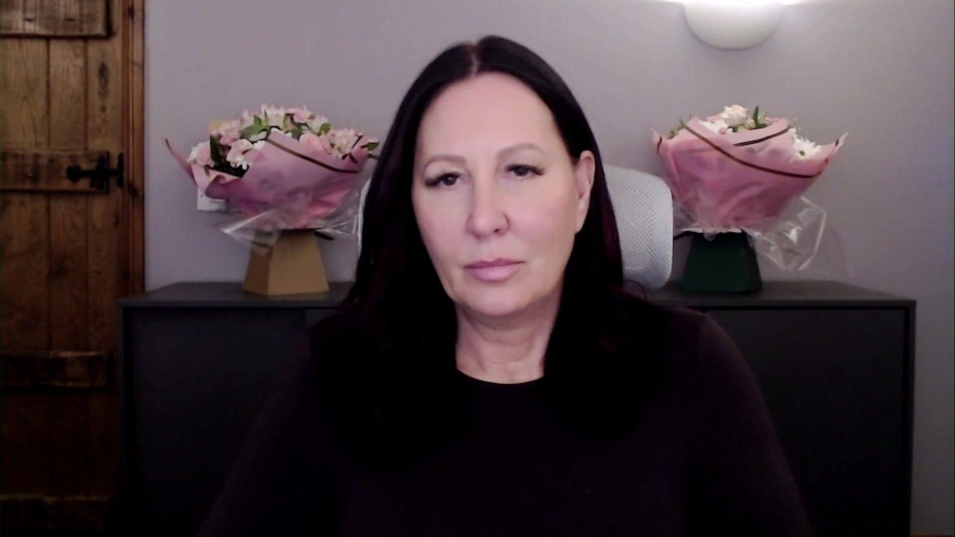 Sasha, who has dark hair and dark clothes, is sitting in the photograph. Behind her are two bouquets of pink flowers. 