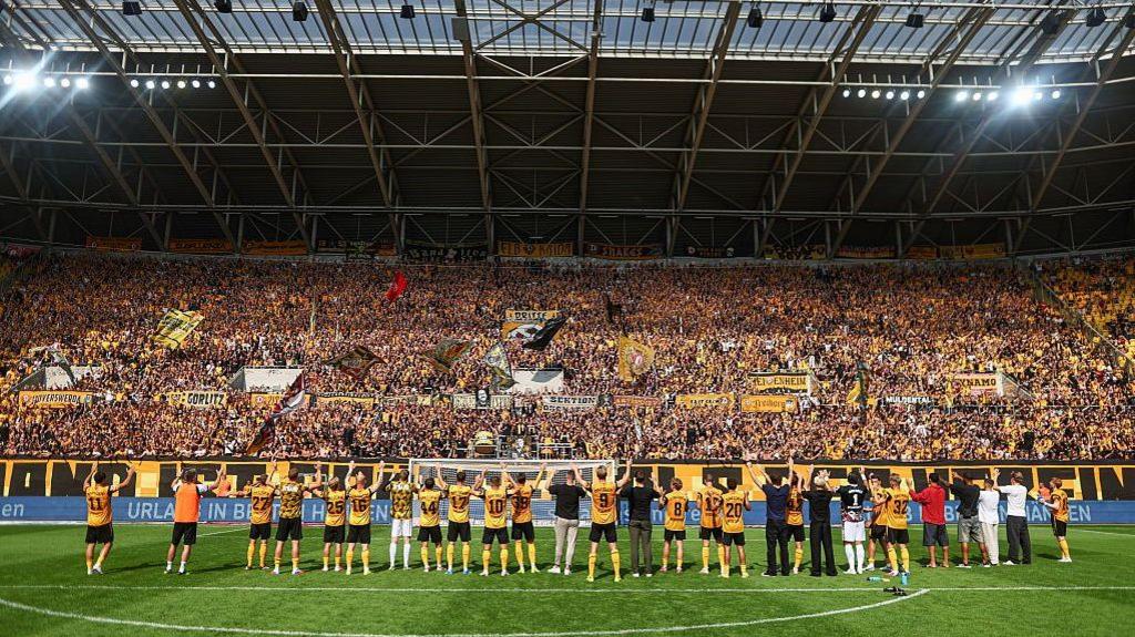 Dynamo Dresden players stand in front of a large, seemingly packed, stand of their own supporters, raising their arms to the fans