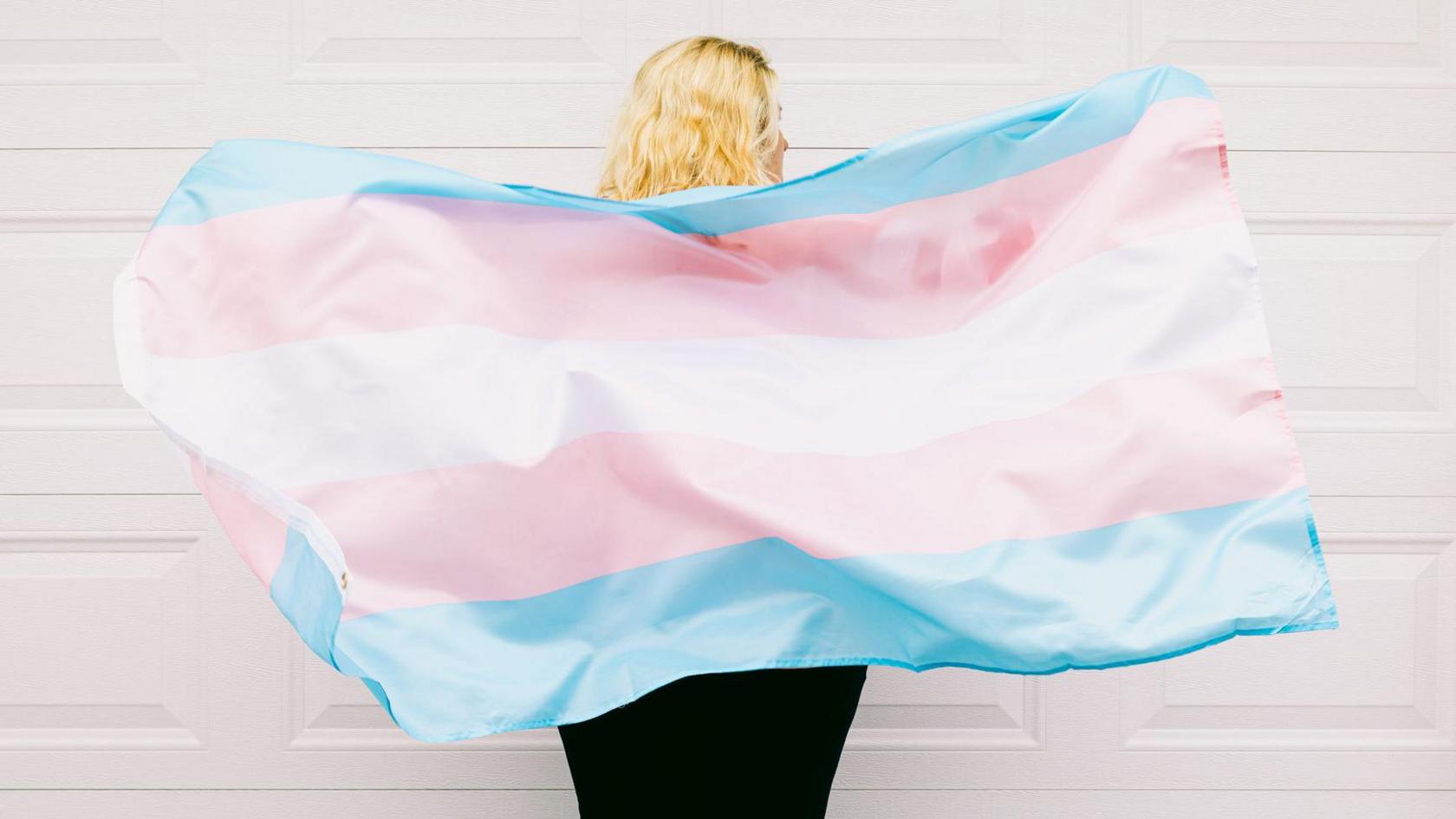 A transgendered person with long blonde hair faces away from the camera and holds the blue, pink and white trans flag behind their back.