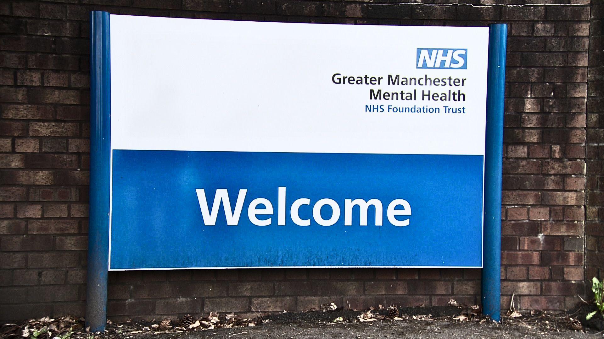 Blue and white NHS signage with WELCOME in white text on a blue background. At the top it says 'Greater Manchester Mental Health NHS Foundation Trust' in black text on white background, below the blue and white NHS logo.