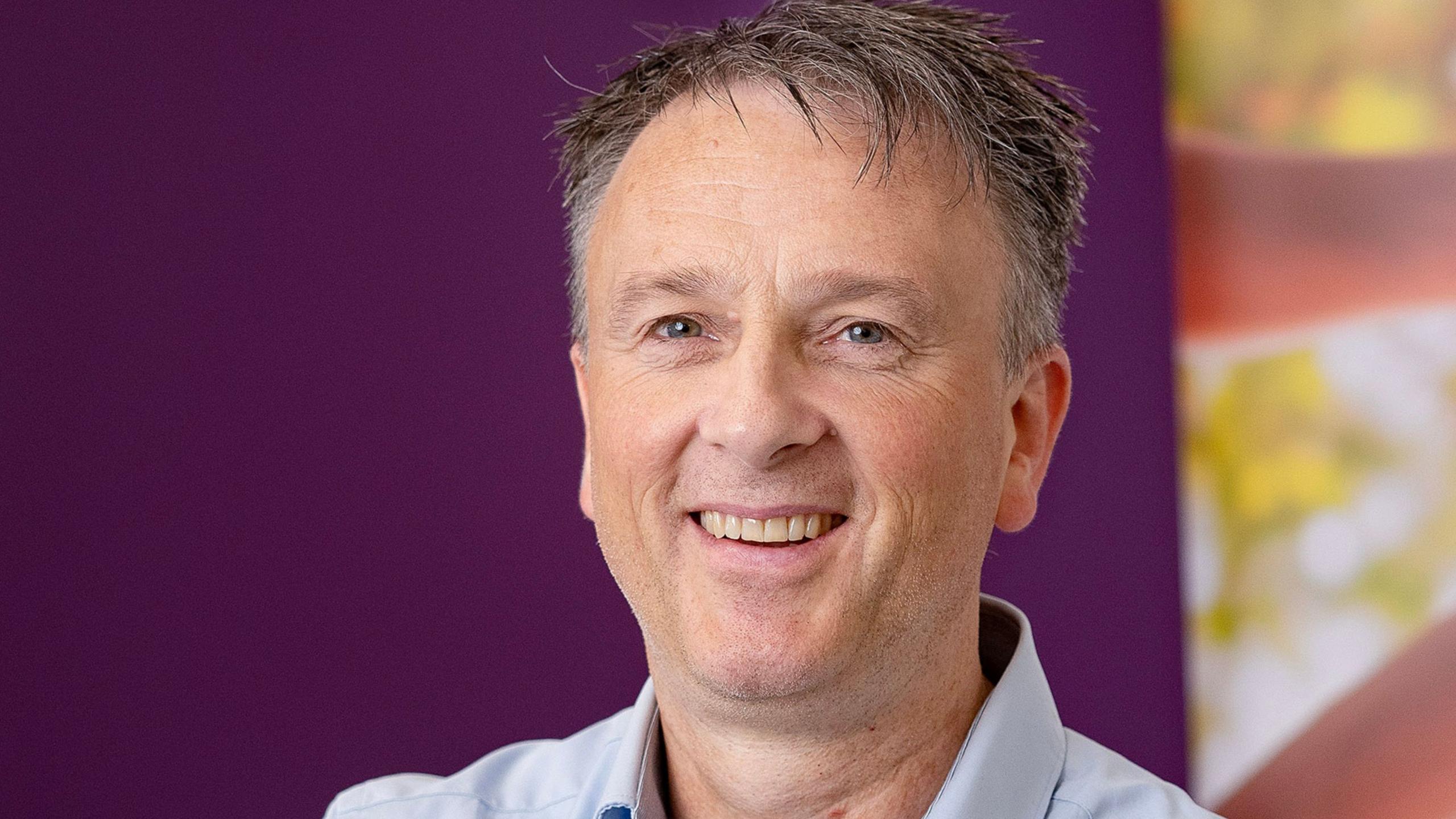 Mal Byrne, a man with short, greying dark hair, smiles at the camera in a charity profile photo. He is wearing a light blue shirt.