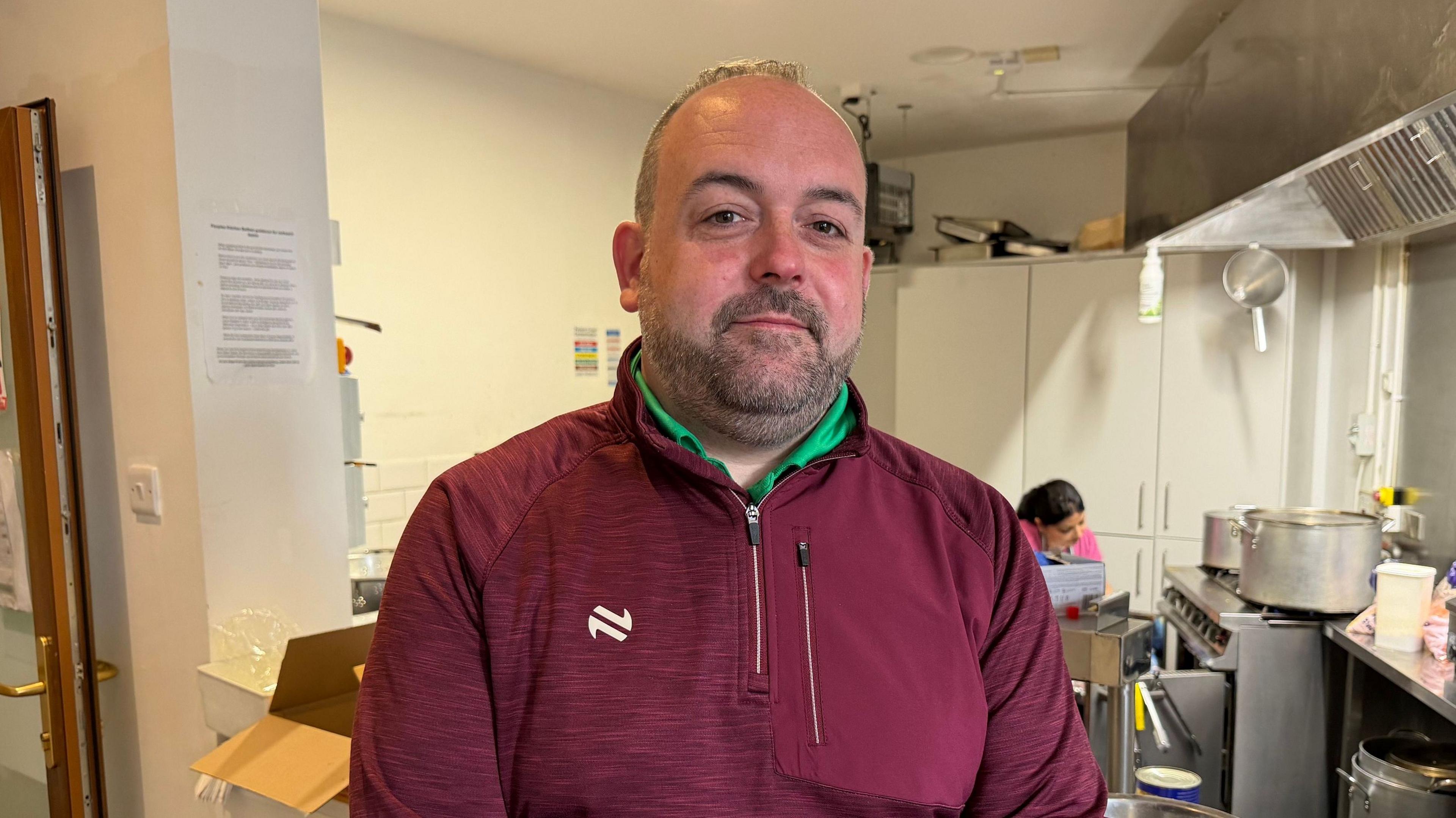 Paul McCusker - a man with short, dark hair and a beard, stands in a soup kitchen. He is wearing a red half-zip top over a bright green shirt. A woman is cooking at a large stove in the background.
