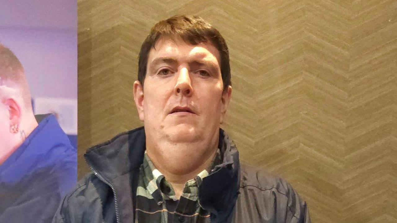 Iain Cameron is wearing a green, orange, black and white chequered shirt with a navy raincoat. He has brown short hair and is looking at the camera.