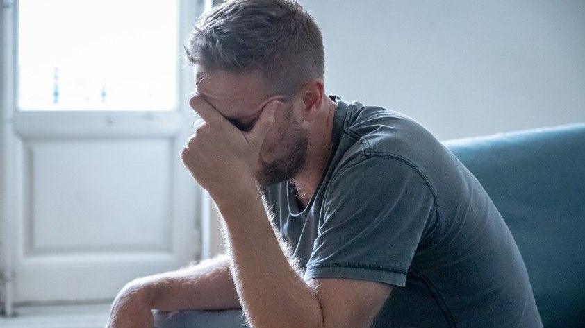 A stock image of a man sat on a sofa with his head in his hands. He is wearing a t-shirt and jeans.