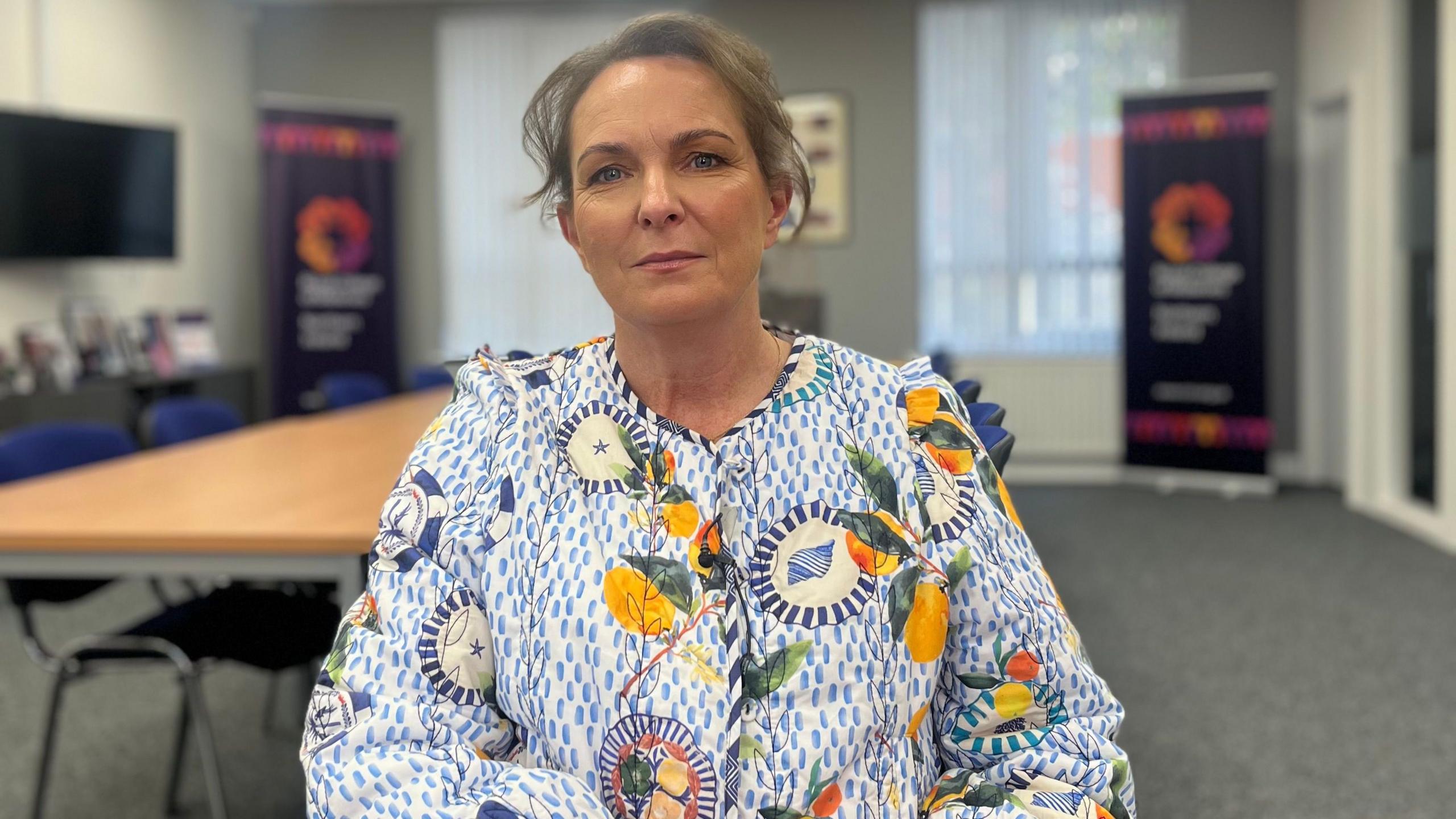 A woman is sitting in a room. She is looking into the camera with a neutral expression. She is wearing a jacket decorated with shells, stars and fruit. Her brown hair is tied up. 
