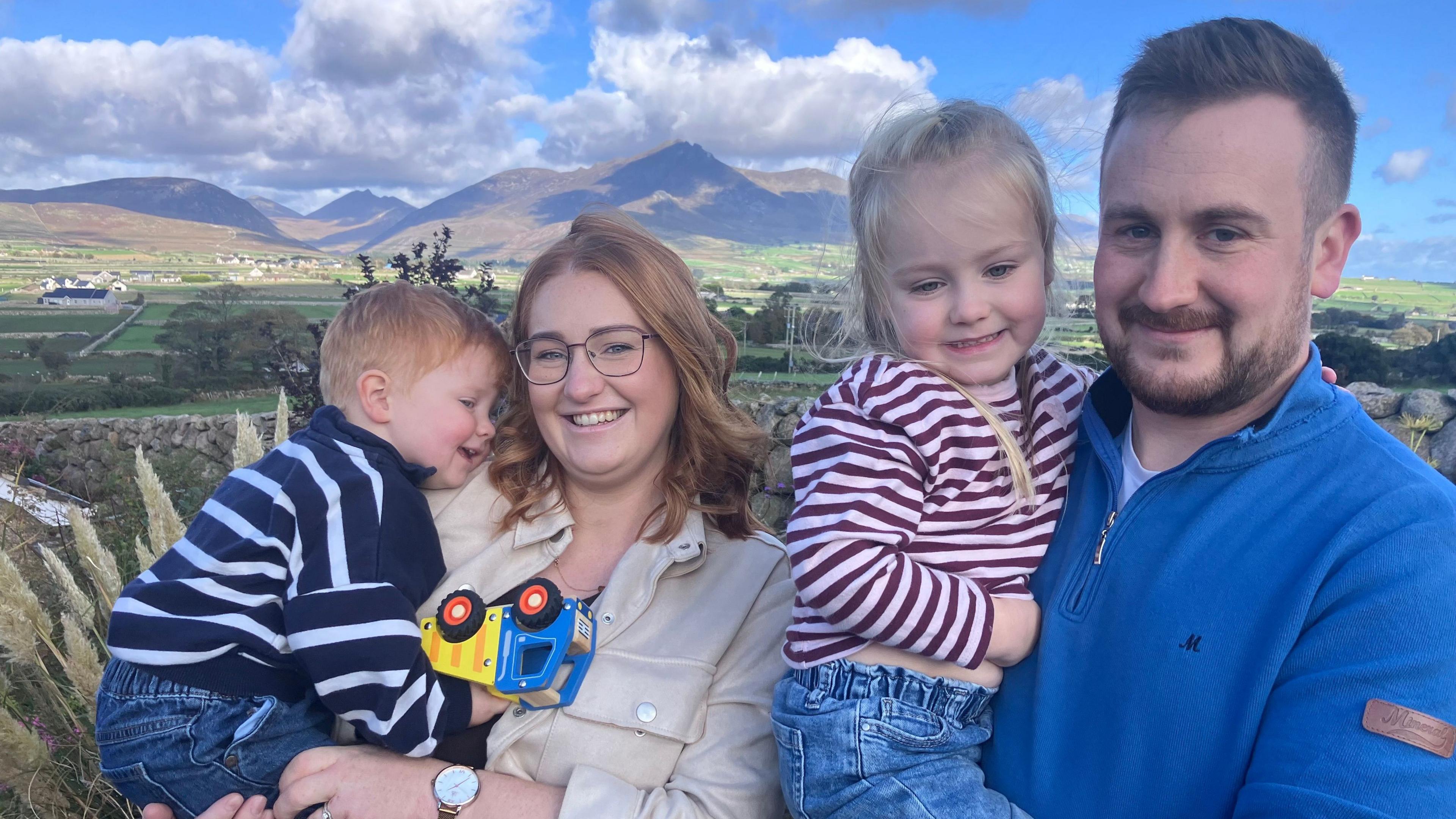 Naomi is holding a little boy. He is holding a toy truck. Mark is holding a little girl. A mountain range and fields are behind them.