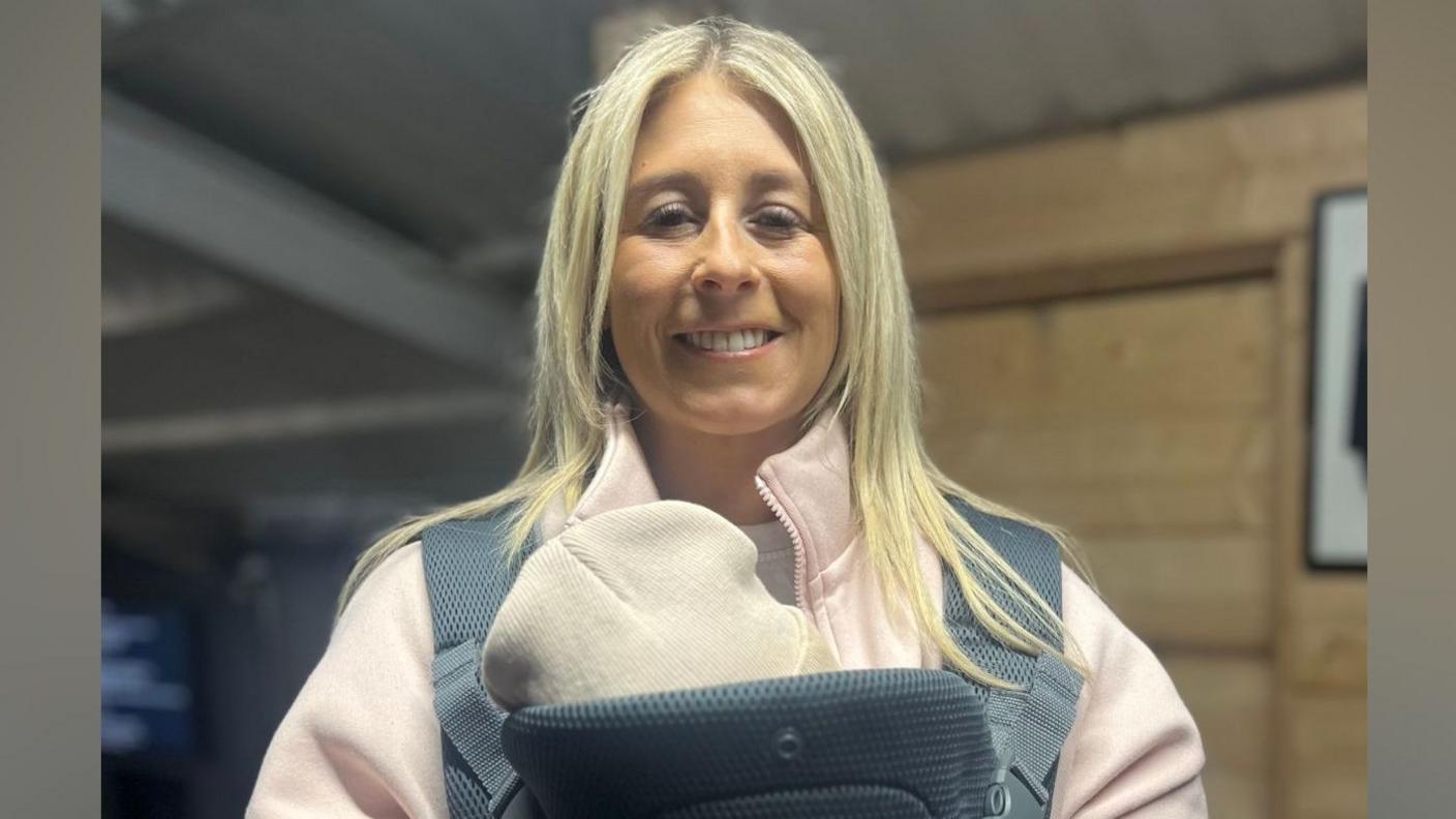 Natasha is standing indoors. A baby is in a harness over her chest. Natasha's blond hair is down. She's wearing a light pink hoodie. Wood panelling is on the walls in the background. 