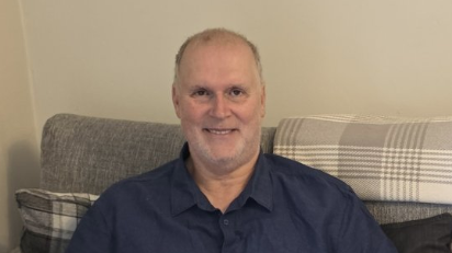 Rex is sitting on a sofa smiling at the camera. He has short grey hair and stubble and is wearing a navy shirt.