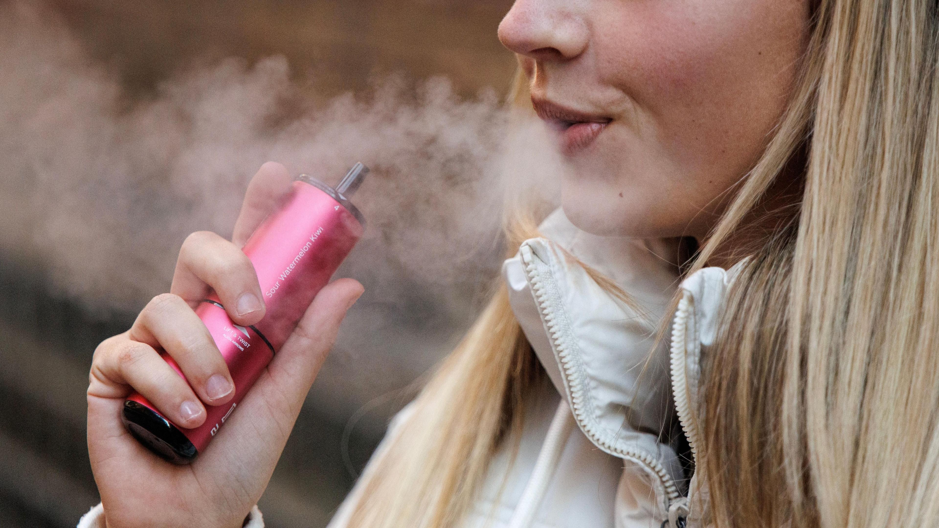A blonde woman breaths out vapour as she holds a red vape in her hand