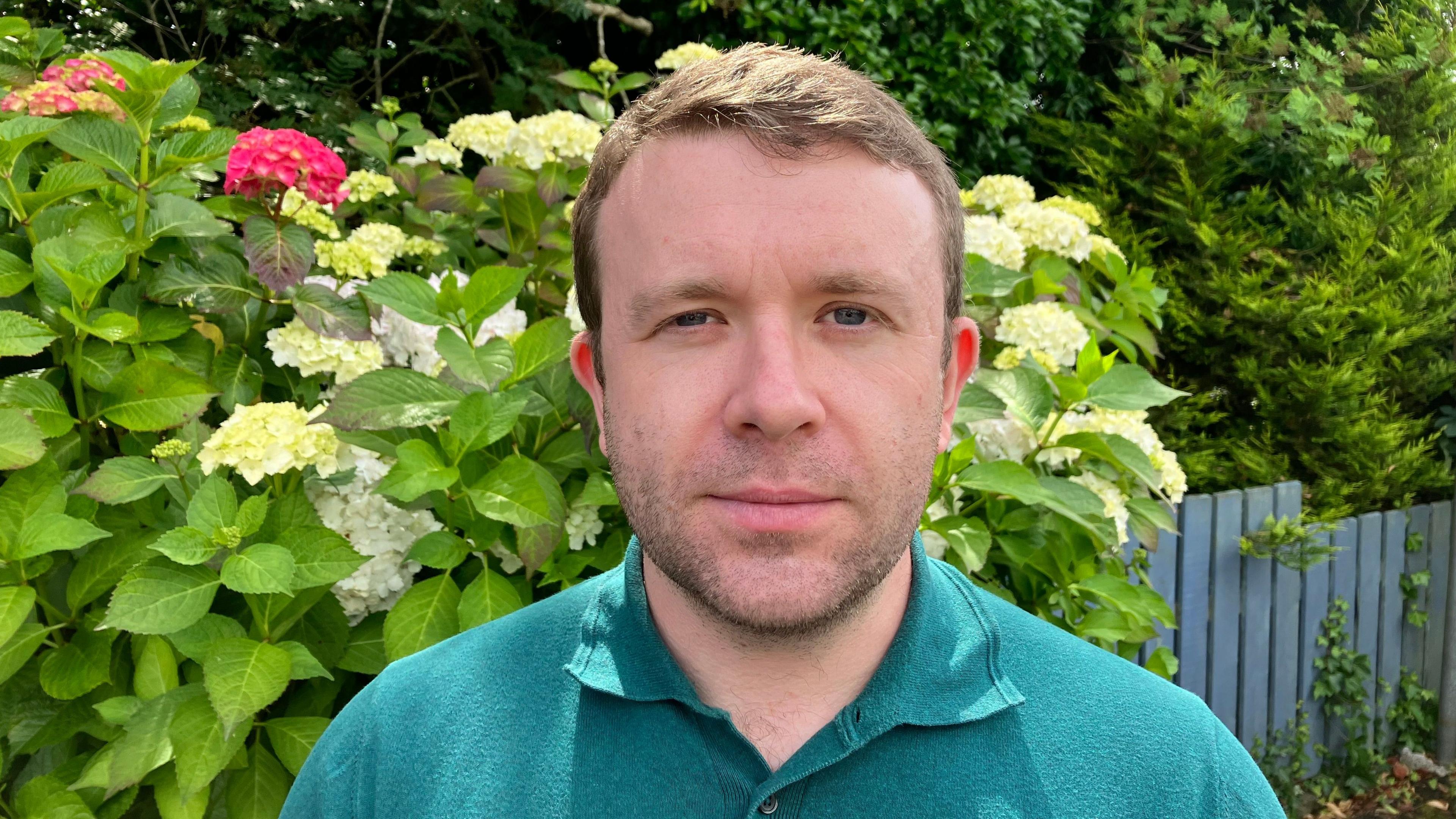A man with brown hair is smiling at a camera. He is wearing turquoise polo top. Behind him is green leaves with pink and white flowers behind him