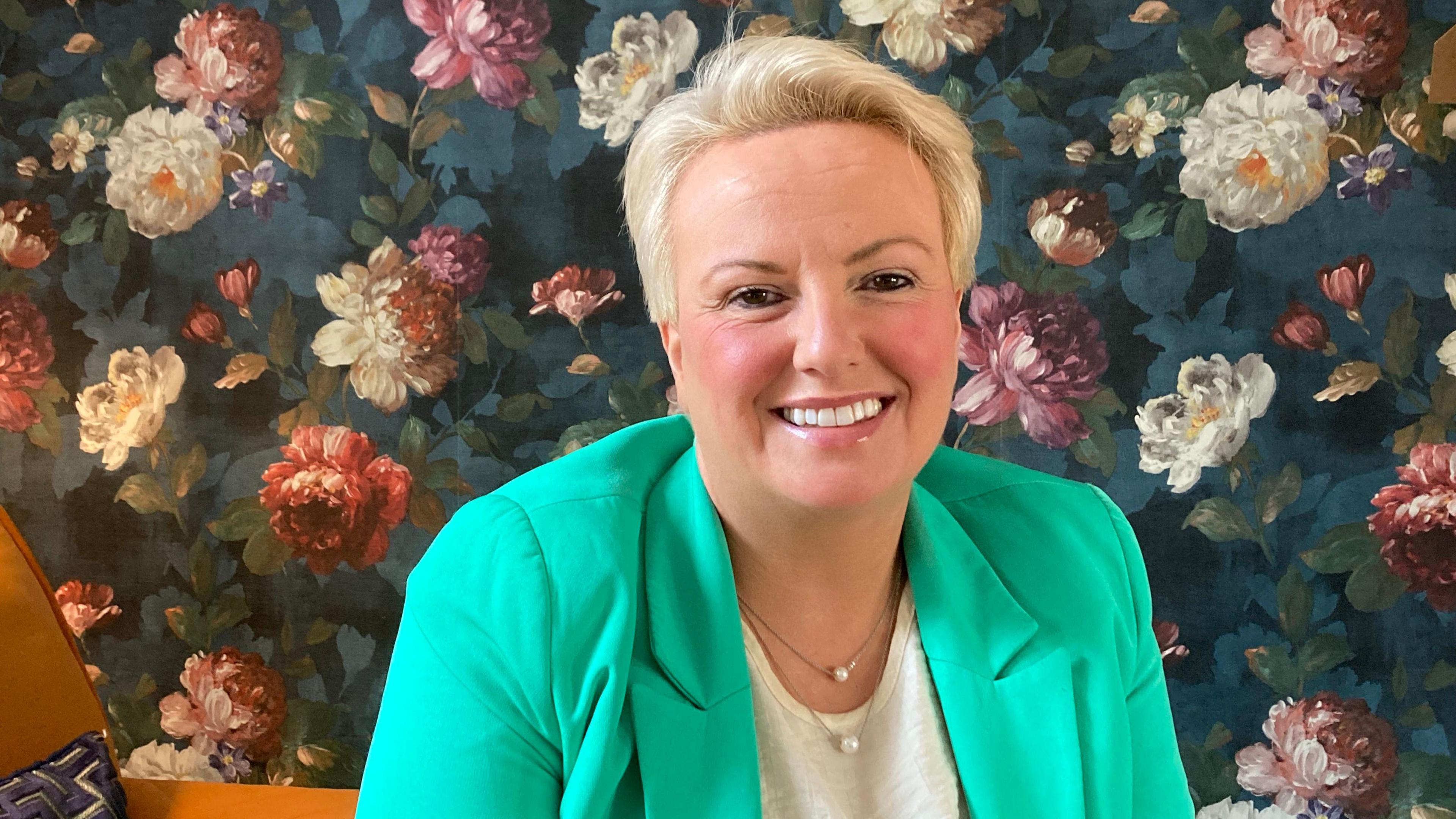 A woman with short blonde hair wearing a green jacket and white top smiling at the camera. She is sitting on an orange armchair in front of wall paper with a flower pattern.