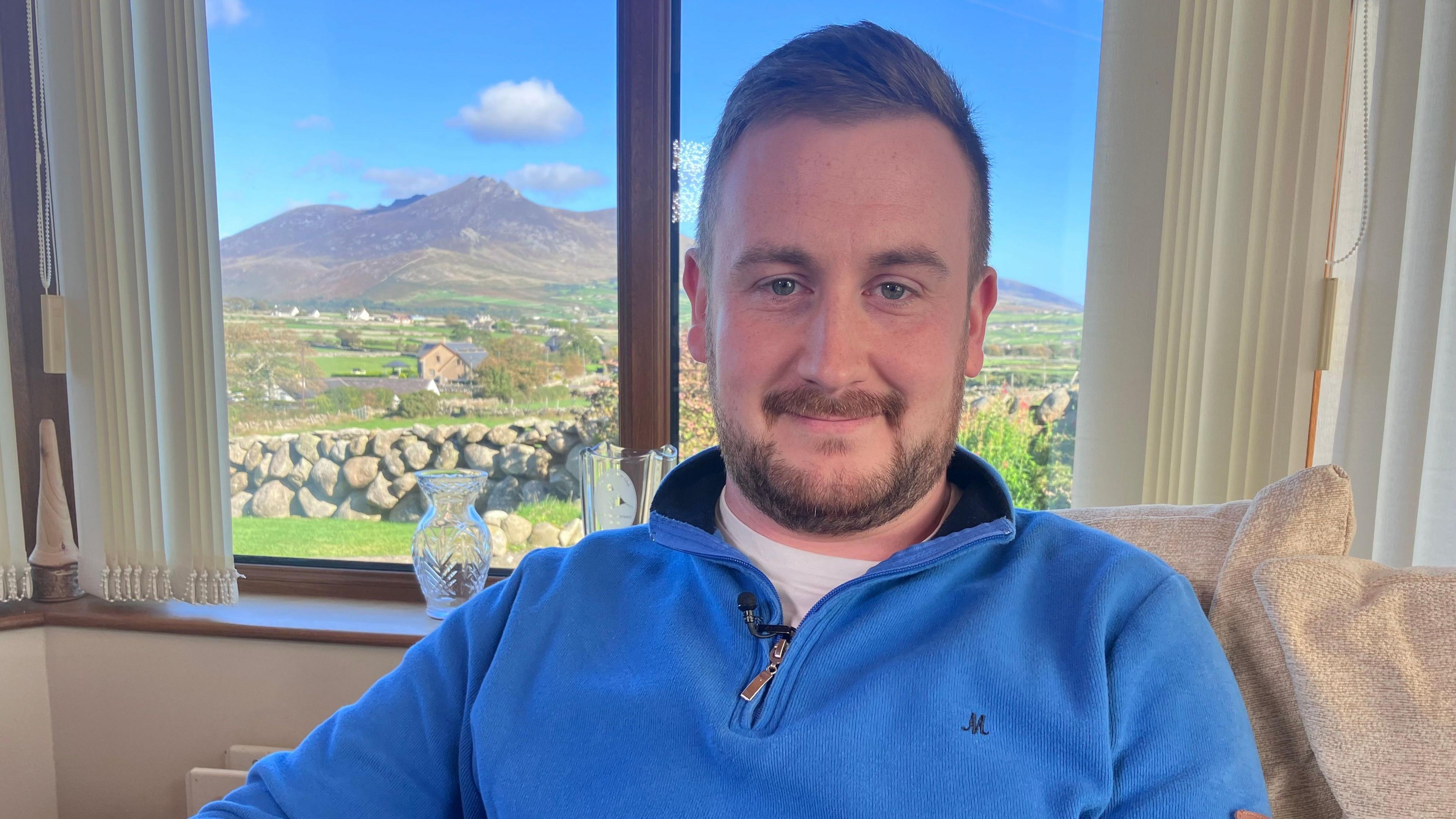 Mark Annett is smiling. He is wearing a blue hoodie. He's sitting on a beige sofa. A crystal vase is on a windowsill. A mountain range is in the background.